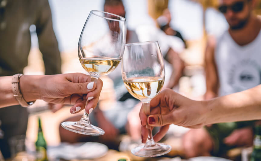 Zwei Personen stoßen mit Weißweingläsern an, der Wein schimmert goldgelb im Licht.